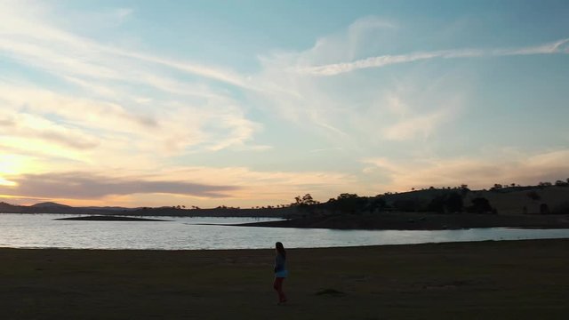 Aerial, orbit, drone shot around a woman, walking on a beach, enjoying the sunset, at lake Huma, on a sunny evening, at Hume Reservoir, at New South Wales and Victoria, in Australia