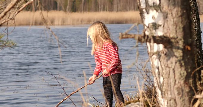 Girl playing alone with big stick by the water the old-fashion way, calm active childhood, contrast to video games