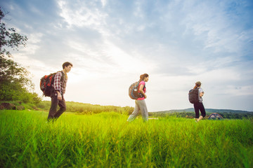 Hikers with backpacks walking through a meadow with lush grass under blue sky and sunset.