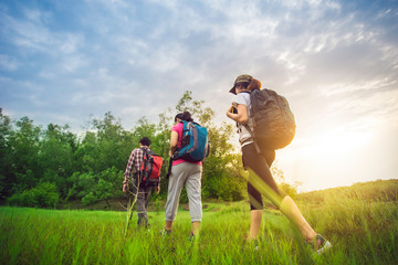 Hikers with backpacks walking through a meadow with lush grass under blue sky and sunset.