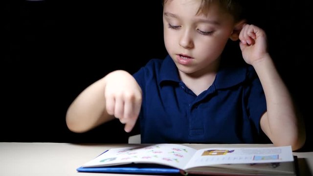 A child sits at night at a table lit by a lamp, reads a book, points his finger at the drawings in the book