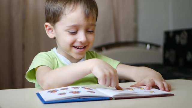 A happy child reading a book with a smile, examines the pictures while sitting at the table at home