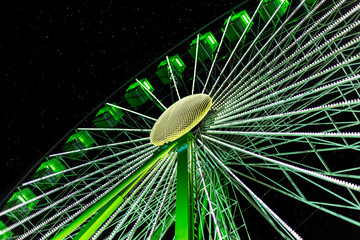 A Ferris wheel turns in the evening in the dark under a starry sky. The wheel is illuminated green and photographed diagonally from below against the sky.