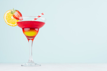 Fresh bright alcohol drink in elegant wineglass with red and yellow liquor, orange slice, strawberry, straw in modern pastel mint interior.