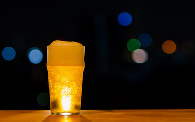 A glass of Beer with its foam puts on wooden table of the bar isolated on dark night background with colorful bokeh lights on rooftop bar.