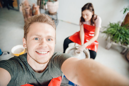 Happy man makes selfie photo on background of girl behind a potter wheel, dishes from clay. Concept loving couple at masterclass of artists - Powered by Adobe
