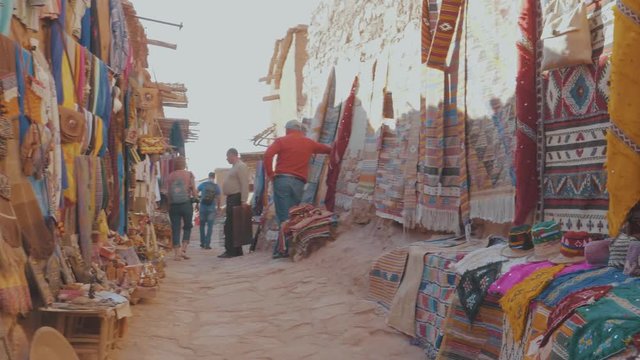 Panning through a narrow street filled with market stalls displaying colorful scarfs bags rugs and other items in an outdoor market place in a Marrakech Souk in Morocco on a sunny day