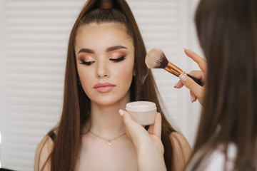 Female makeup master use a powder brush. Beautiful model in beauty studio. Professional make up artist finish fashion maleup. Background of white folding screen
