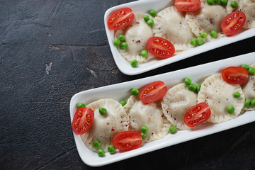 Dumplings with green peas and cherry tomatoes served on two white plates, closeup over dark brown stone surface, elevated view