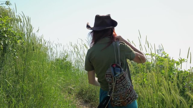 A young red-haired traveler girl in a cowboy hat with a backpack walks along a mountain path overgrown with grass and shrubs.