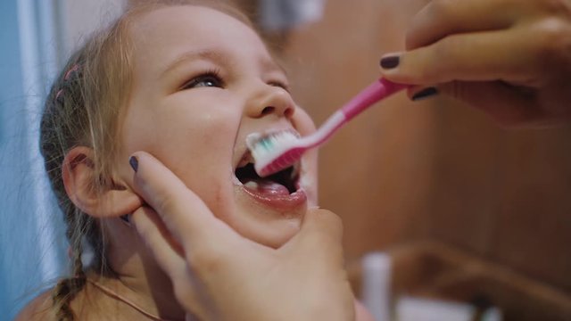 Mother helping her cute european appearance daughter brush her teeth at home bathroom, mom helps to clean the teeth to her daughter. Close-up, slow motion