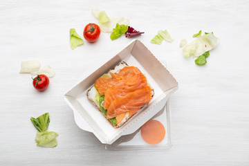 Top view sandwich with soft cheese and red fish lies in the lunch box next to the greens and tomatoes on a white table. Concept of a healthy snack.