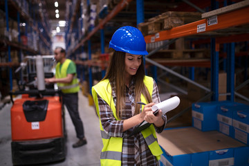 Female warehouse worker checking inventory in storage area while her coworker driving forklift in...