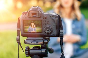 Stylish young girl blogger sitting on the grass in front of camera and shoots video. Young blonde woman working as a blogger recording video tutorial