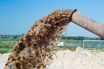 The flow of brown liquid from the pipe close-up against the blue clear sky and green plants.