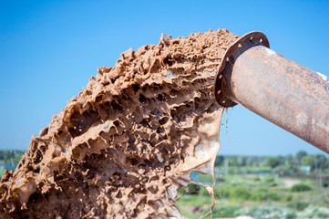 The flow of brown liquid from the pipe close-up against the blue clear sky and green plants.