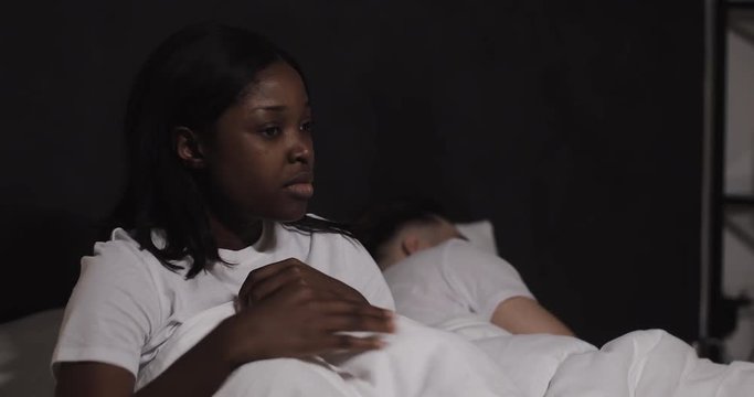 Married multi-ethnic couple lying in bed at night. Worried woman sitting on bed feels sad, pensive wife looking at sleeping husband, difficulties in relationships, thinking of breaking up divorce.