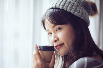 Happy Asian woman wearing sweater and hat, holding coffee mug near window at home.
