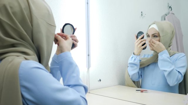 Muslim young attractive woman in beige hijab and traditional blue dress making make-up and standing in front of the mirror.