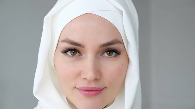 Portrait of young attractive muslim woman wearing white hijab is rising her eyes, looking at the camera and smiling on light background.