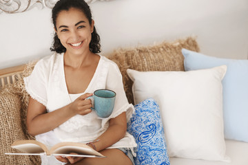 Obraz na płótnie Canvas Happy relaxed good-looking tanned brunette woman enjoy weekend day-off stay home indoors sitting comfortable rattan couch living room look camera smiling broadly hold coffee mug reading book