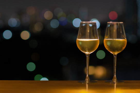 Two glasses of white wine on wooden table of rooftop bar with colorful bokeh of city light and space for text.