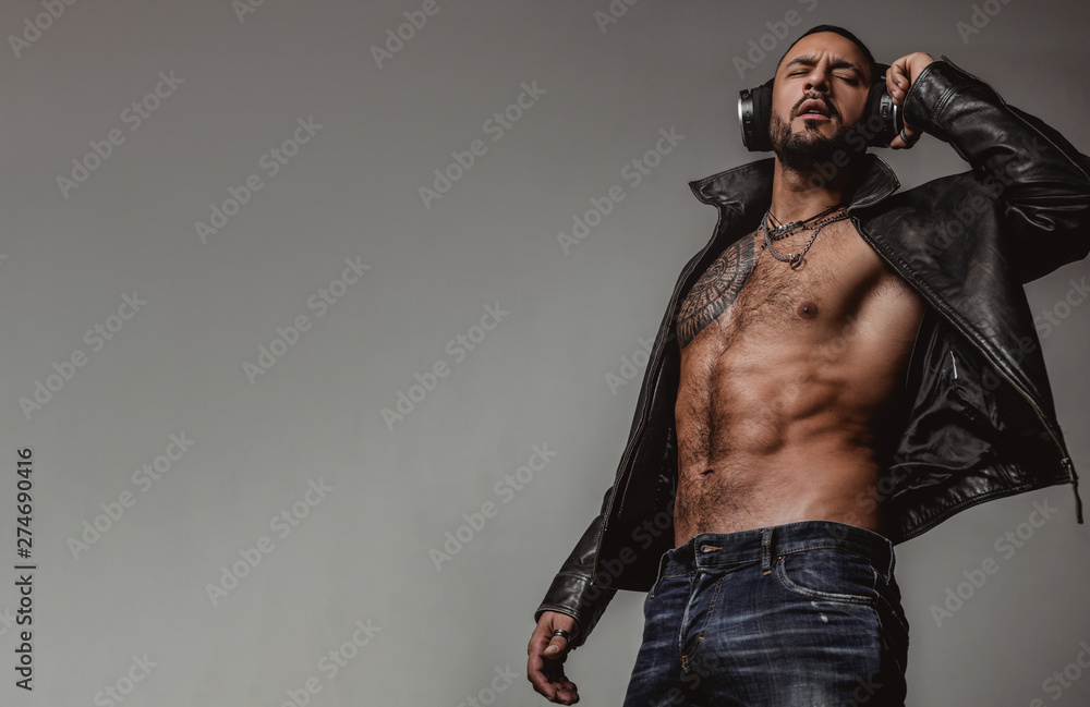 Wall mural Music for Dj concept. Hot brutal man with a beard, a tattoo wearing jeans and black leather jacket. Attractive model enjoys music in headphones.