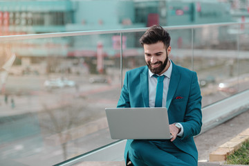 Young successful smiling bearded businessman in blue suit sitting on rooftop and using laptop.