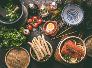 Healthy ingredients for low carb meal cooking: salmon in asian bamboo steamer , asparagus bunch, fresh herbs and oil and sauce bottles on dark table background, top view. Asian cuisine. Keto diet