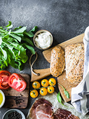 Various fresh ingredients for making sandwiches - vegetables, bread, prosciutto, olive oil and curd cheese on dark background. Top view. copy space