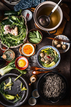 Top view of Asian food dishes. Asian noodle soup preparation on dark rustic kitchen table. Pot with stock, cooked noodles, various vegetables and meatballs. Meatball noodle soup in bowl with chopstick