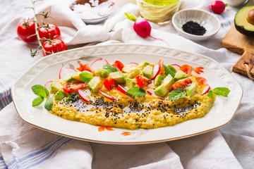 Close up of hummus plate topped with olive oil, avocado, fresh vegetables and herbs served on kitchen table, top view. Vegan food concept. Healthy snack. Mediterranean lunch.