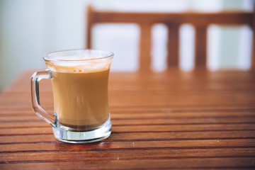 Glass or cup of coffee with white foam on brown wooden table on the balcony, with wooden chair in the background, with copy space
