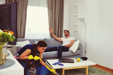 couple relationship concept, marriage  living problems and conflict, young couple in living room woman cleaning and vacuuming house, man sitting on sofa watching television, home jobs gender equality 