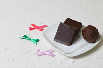 Chocolate cake pops and pieces of black chocolate on a white plate. Near bows for decoration. On the surface covered with white decorative plaster.