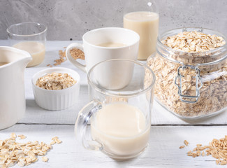 Close-up set of glasses and bottle with oat milk, jar with oat flakes on white wooden table near concrete wall. 