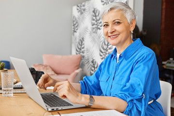 Side view of successful mature female blogger sitting at home office with digital camera and open...