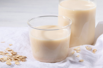 Close-up glass of oat milk and flakes on white linen napkin on white table.
