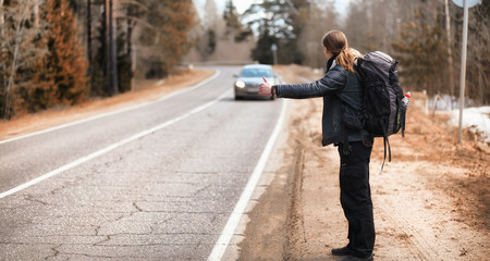 A young man is hitchhiking around the country. The man is trying to catch a passing car for traveling. The man with the backpack went hitchhiking to the south.