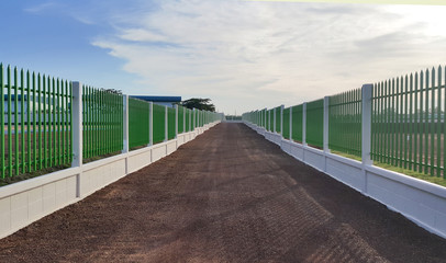 The gravel road lies between the white cement fence with green iron bars under the blue sky.