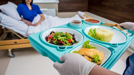 Nurse in medical coat is holding a tray with breakfast for the young female patient.