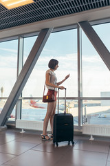 Young casual female traveler in airport, holding smart phone near gate windows at planes on runway.