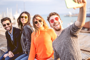 Young group of friends taking selfies sitting on a harbor wall wearing sunglasses. Youth having fun using new technologies concept.
