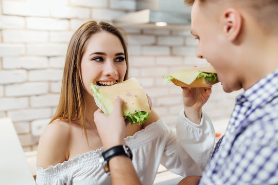 Husband reaching over his wife's shoulder to taste the sandwich which she is offering him. - Powered by Adobe