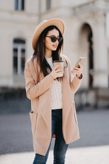 Obraz premium Cheerful young woman wearing beige coat using her phone in the sunny city street and drinking take away coffee in paper cup.