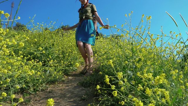 A young red-haired traveler girl in a cowboy hat walks along a mountain path overgrown with grass and shrubs with yellow flowers.