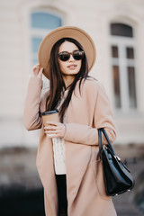 Young woman wearing coat and black hat walking in city street and drinking take away coffee in...