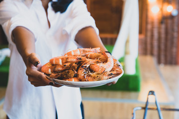 Close-up grilled prawn on dish hold by tourist woman. Group of tourist having BBQ, Seafood party at seaside...