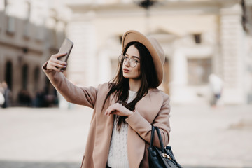 Young attractive playful tourist is making selfie on the phone outside, wearing hat, sunglasses, coat and handbag