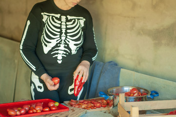 Young female cook in skeleton costume and apron cuts red tomatoes for making tomato sauce. Hands close up.